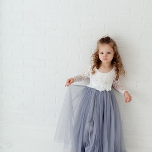 May include: A young girl wearing a white lace bodice and a long, flowing grey tulle skirt. She is standing in front of a white brick wall.