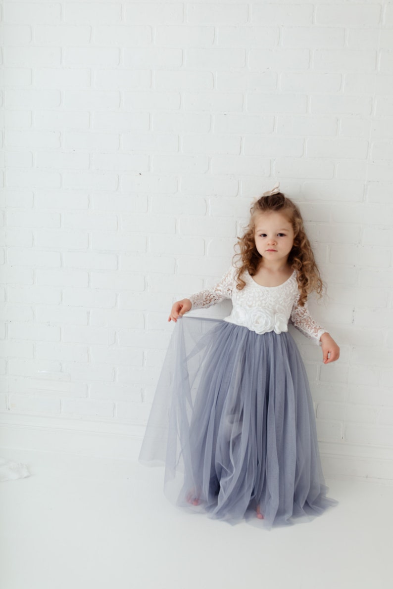 May include: A young girl wearing a white lace bodice and a long, flowing grey tulle skirt. She is standing in front of a white brick wall.