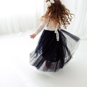 May include: A young girl wearing a white lace top and a long, flowing black tulle skirt. She has long, curly brown hair with white bows in it. She is twirling in a white room.