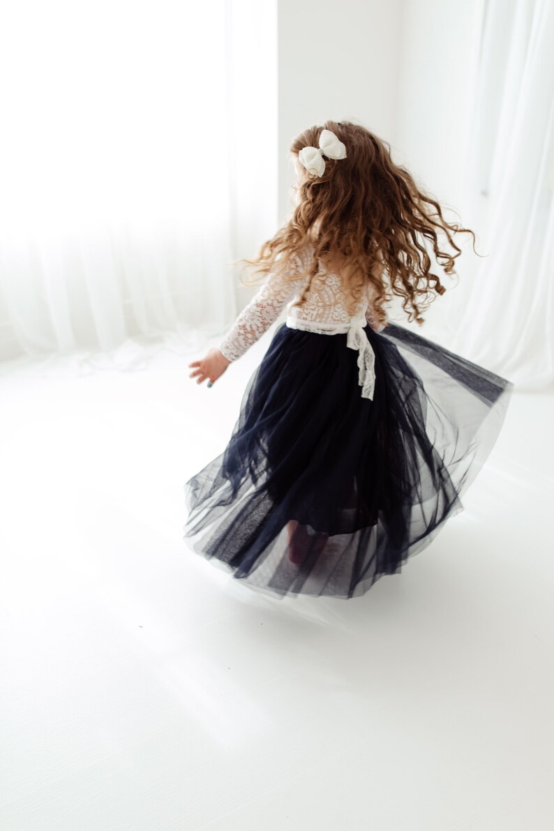 May include: A young girl wearing a white lace top and a long, flowing black tulle skirt. She has long, curly brown hair with white bows in it. She is twirling in a white room.