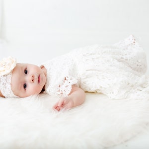 K&ouml;nnte beinhalten: Ein Baby M&auml;dchen in einem wei&szlig;en Spitzenkleid und einem Stirnband mit einer wei&szlig;en Blume. Das Baby liegt auf einer wei&szlig;en, flauschigen Oberfl&auml;che.