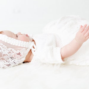 May include: A white lace bonnet with a delicate floral pattern and a matching white lace dress. The bonnet features a scalloped edge and a tie closure under the chin. The dress has short sleeves and is crafted from a sheer, intricate lace fabric.