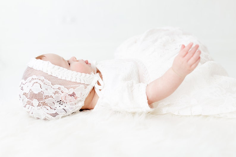 May include: A white lace bonnet with a delicate floral pattern and a matching white lace dress. The bonnet features a scalloped edge and a tie closure under the chin. The dress has short sleeves and is crafted from a sheer, intricate lace fabric.