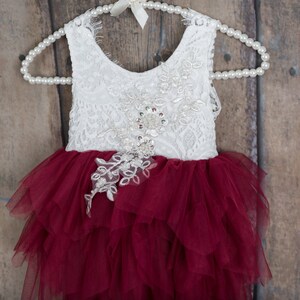 Puede incluir: Vestido de encaje blanco con falda de tul burdeos, colgado de una percha de perlas. El corpi&ntilde;o presenta adornos florales con lentejuelas y abalorios. La falda tiene m&uacute;ltiples capas de tul con volantes.