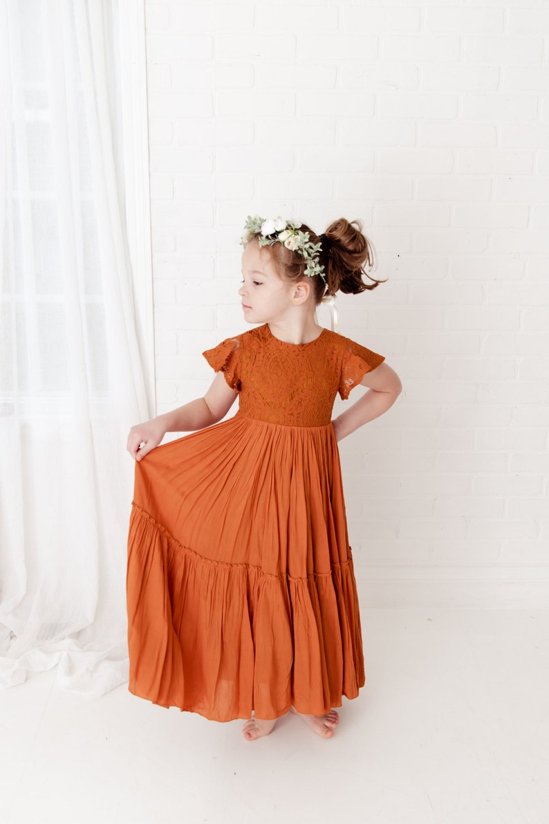 Op de afbeelding: Een jong meisje draagt een lange, roestkleurige jurk met kantdetails en een bloemenkrans. De jurk heeft een gelaagde rok en korte mouwen.