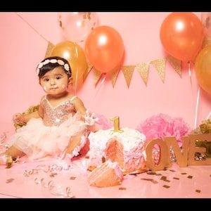 May include: A baby girl in a pink tutu and sequined top sits in front of a pink cake decorated with pink frosting and a number one. The cake is surrounded by gold confetti and balloons. The background is pink with a gold glitter banner that says "LOVE".