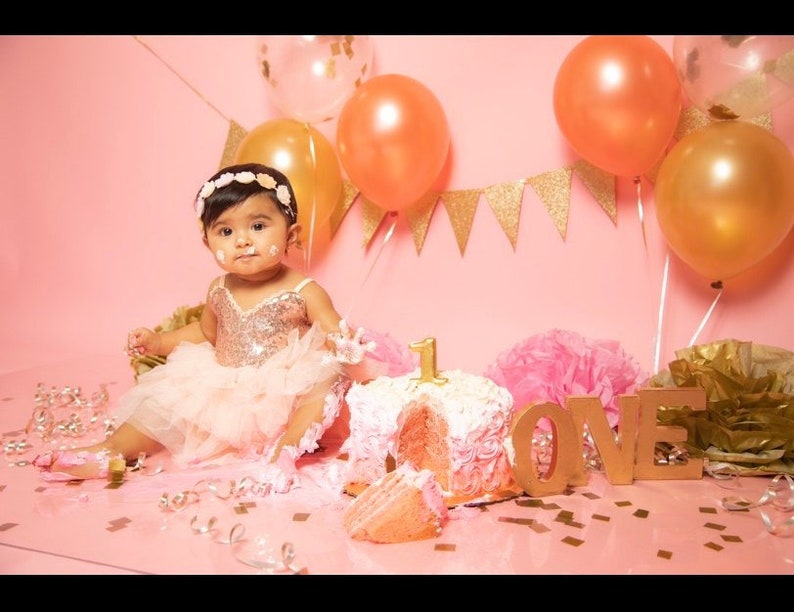 May include: A baby girl in a pink tutu and sequined top sits in front of a pink cake decorated with pink frosting and a number one. The cake is surrounded by gold confetti and balloons. The background is pink with a gold glitter banner that says "LOVE".