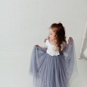 May include: A young girl wearing a white lace bodice and a long, flowing grey tulle skirt. She is holding the skirt up with her hands and looking to the side.