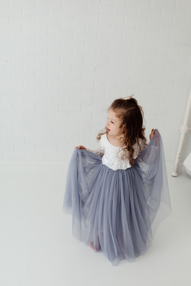 May include: A young girl wearing a white lace bodice and a long, flowing grey tulle skirt. She is holding the skirt up with her hands and looking to the side.