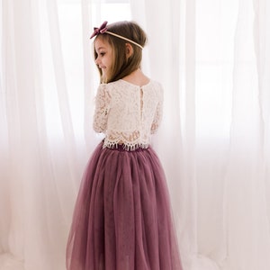 May include: A young girl wearing a white lace long-sleeved top and a long, dusty rose tulle skirt. She is wearing a burgundy bow headband.