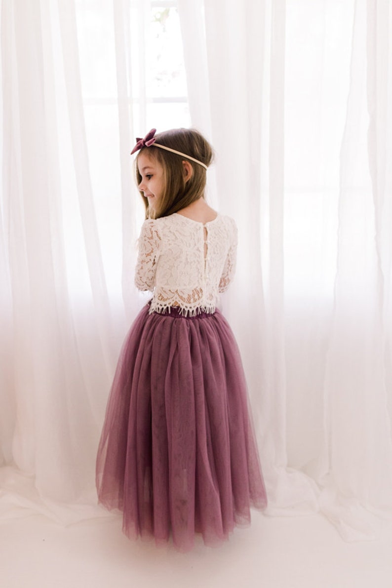 May include: A young girl wearing a white lace long-sleeved top and a long, dusty rose tulle skirt. She is wearing a burgundy bow headband.