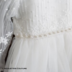 K&ouml;nnte beinhalten: Wei&szlig;es T&uuml;llkleid mit Perlenbesatz und Spitzendetails. Das Kleid hat lange &Auml;rmel und ein tailliertes Oberteil.
