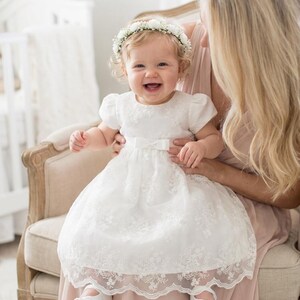 May include: A white infant dress featuring short puff sleeves, a round neckline, and a floral lace overlay with a scalloped hem. The bodice is accented with a small center bow at the waist. The outfit is paired with white soft-soled shoes.