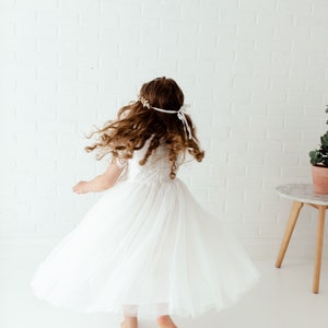 May include: A young girl wearing a white tulle dress with lace detailing, spinning around in a white room. She has long brown hair and is wearing a floral headband.