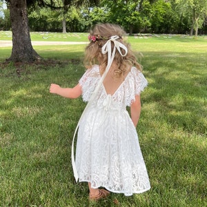 May include: A young girl wearing a white lace dress with a flower crown and a white ribbon tied in a bow at the back of her neck. She is standing in a grassy field.