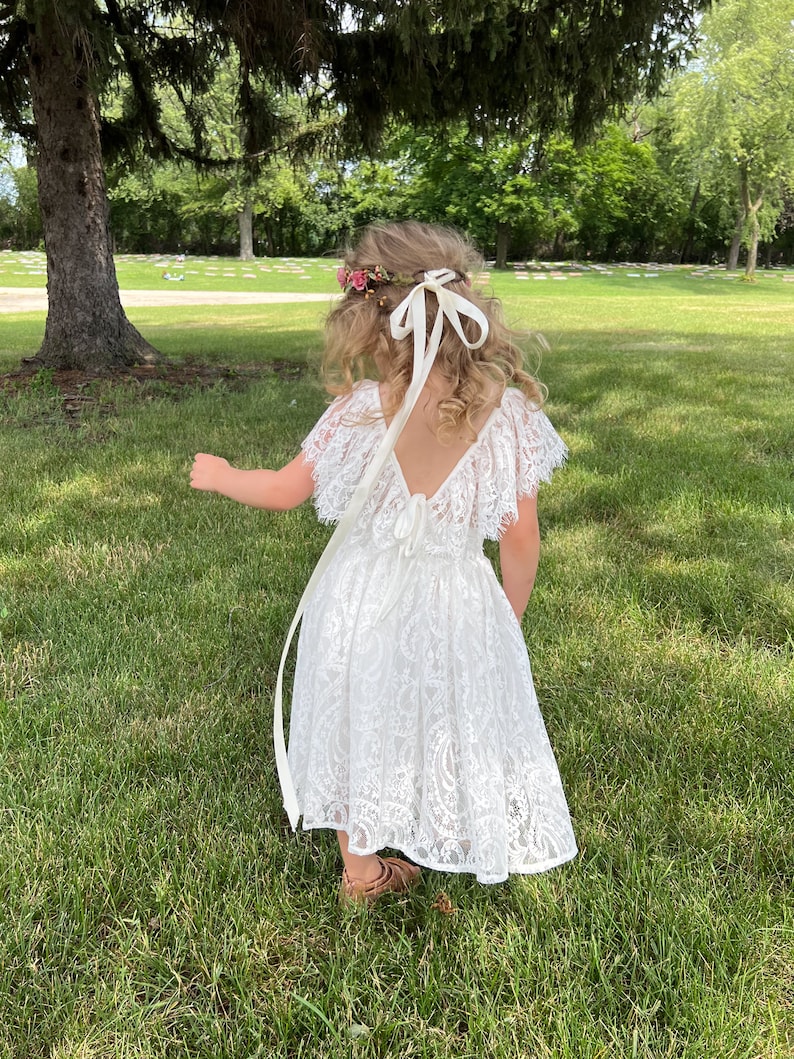 May include: A young girl wearing a white lace dress with a flower crown and a white ribbon tied in a bow at the back of her neck. She is standing in a grassy field.
