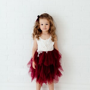 Puede incluir: Una ni&ntilde;a peque&ntilde;a con un vestido blanco y burdeos con una falda de tul en capas. El corpi&ntilde;o es blanco con detalles intrincados. Tiene el pelo rizado y un lazo burdeos. El fondo es una pared de ladrillos blancos.