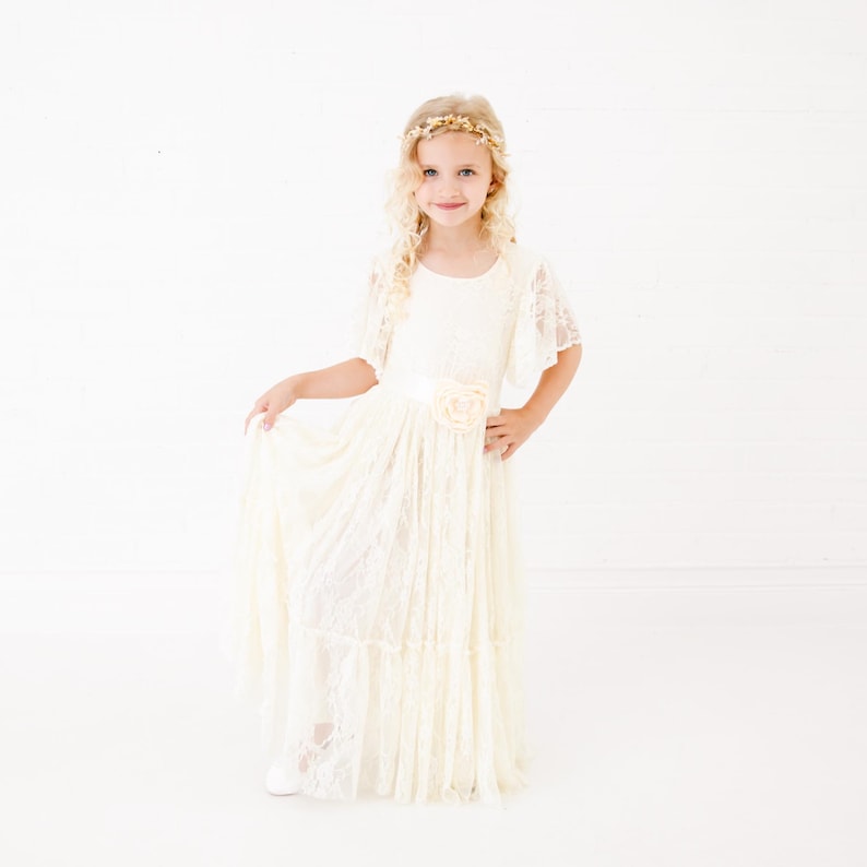 Peut inclure: Une jeune fille portant une robe en dentelle blanche avec un accent floral &agrave; la taille. Elle porte une couronne de fleurs dor&eacute;es et a les cheveux blonds boucl&eacute;s.