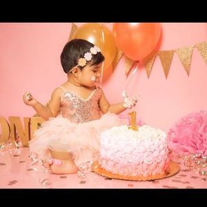May include: A young girl in a pink and gold sequined dress with a tulle skirt is celebrating her first birthday. She is wearing a flower headband and is holding a piece of cake with icing on her fingers. The cake is decorated with pink and white icing and a gold number one on top. There are pink and gold decorations in the background.