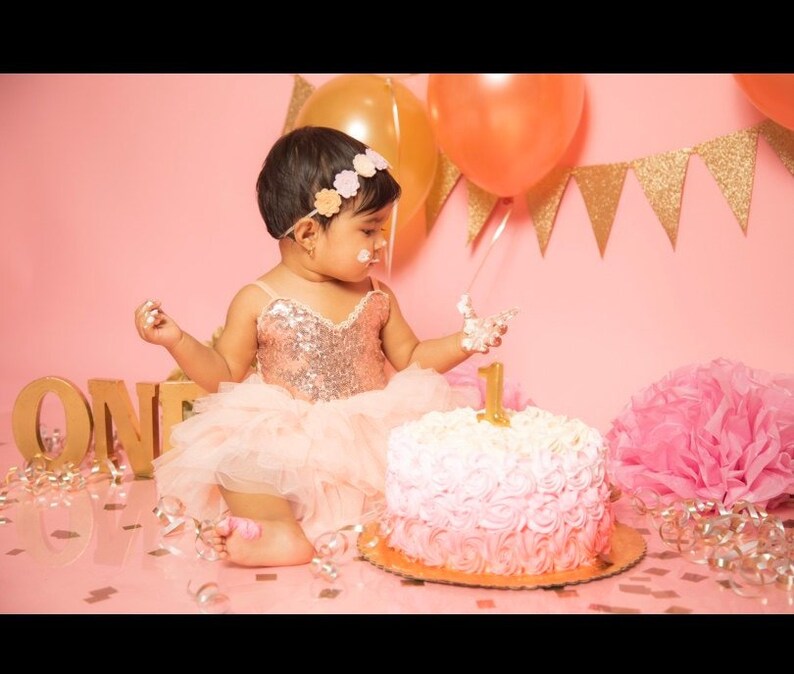 May include: A young girl in a pink and gold sequined dress with a tulle skirt is celebrating her first birthday. She is wearing a flower headband and is holding a piece of cake with icing on her fingers. The cake is decorated with pink and white icing and a gold number one on top. There are pink and gold decorations in the background.