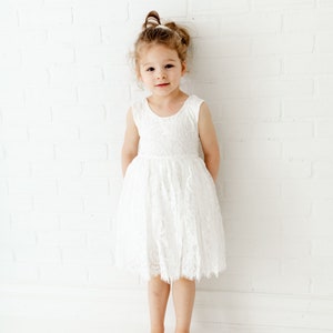 Pode incluir: Uma menina pequena vestindo um vestido branco de renda com uma faixa de cabe&ccedil;a branca. Ela est&aacute; em p&eacute; em frente a uma parede de tijolo branco.
