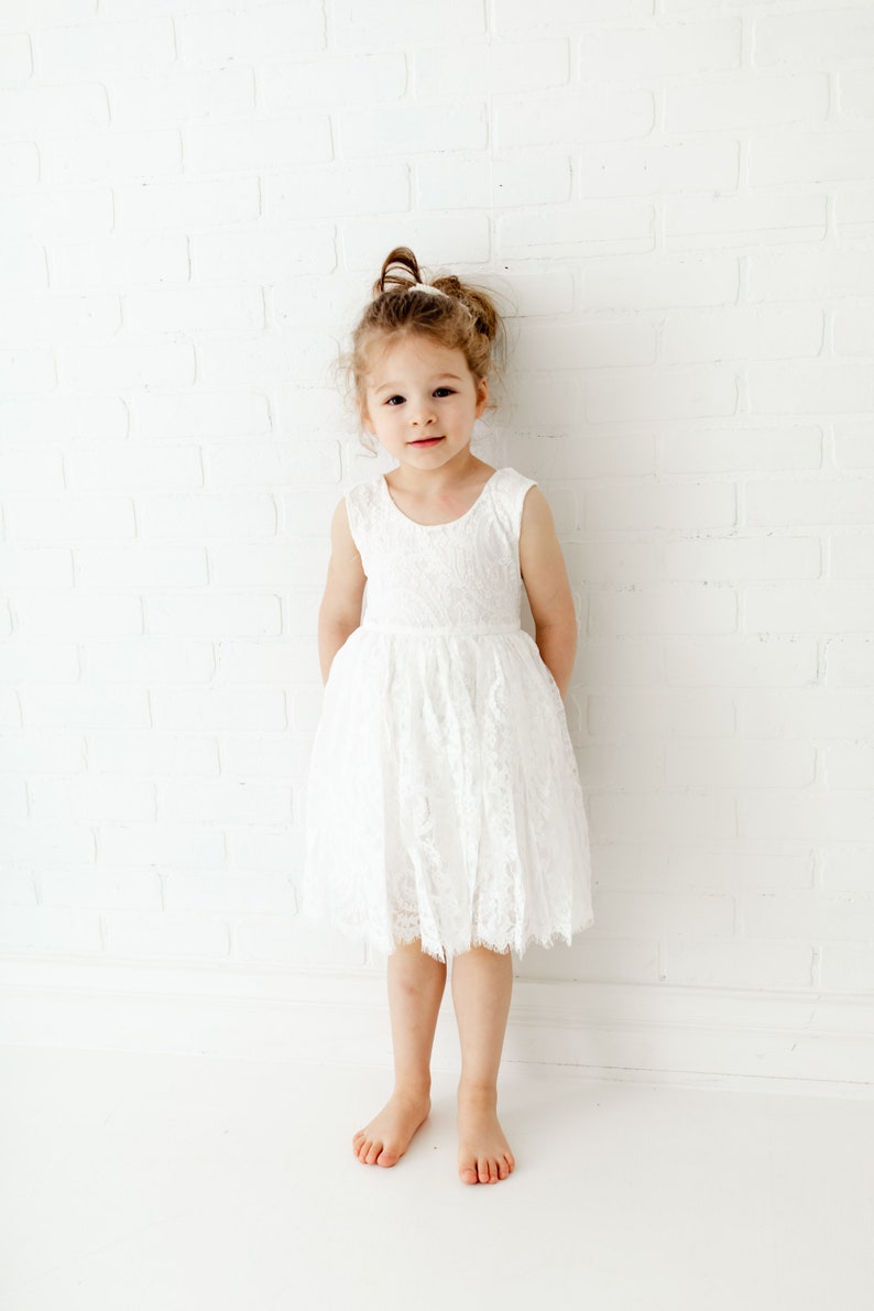Pode incluir: Uma menina pequena vestindo um vestido branco de renda com uma faixa de cabe&ccedil;a branca. Ela est&aacute; em p&eacute; em frente a uma parede de tijolo branco.