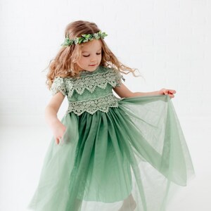 May include: A young girl wearing a sage green tulle dress with lace detailing and a leafy green crown. The dress has a full skirt and the girl is twirling, showing off the layers of tulle.