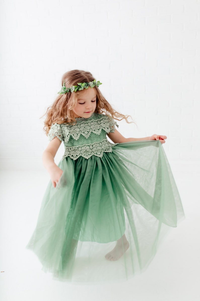 May include: A young girl wearing a sage green tulle dress with lace detailing and a leafy green crown. The dress has a full skirt and the girl is twirling, showing off the layers of tulle.