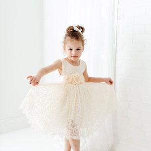 May include: A young girl wearing a white lace dress with a flower embellishment on the waist. She is standing on a white floor and smiling at the camera.