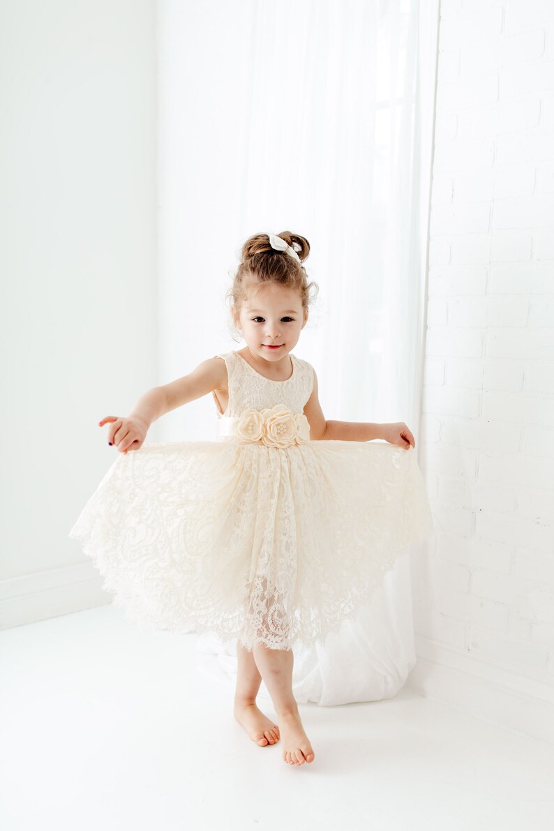 May include: A young girl wearing a white lace dress with a flower embellishment on the waist. She is standing on a white floor and smiling at the camera.