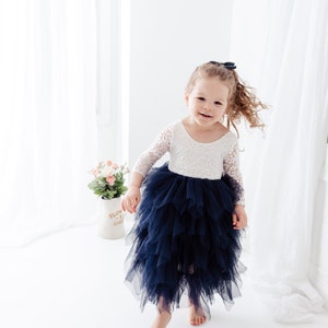 May include: A dress featuring a white lace bodice with three-quarter length sleeves and a voluminous, layered navy blue tulle skirt. The garment has a scoop neckline and a fitted waistline, with the skirt falling to a mid-calf length.