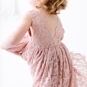 Pu&ograve; includere: Una bambina che indossa un abito di pizzo rosa polvere con schiena a V. L'abito ha una gonna fluida ed &egrave; perfetto per un'occasione speciale.