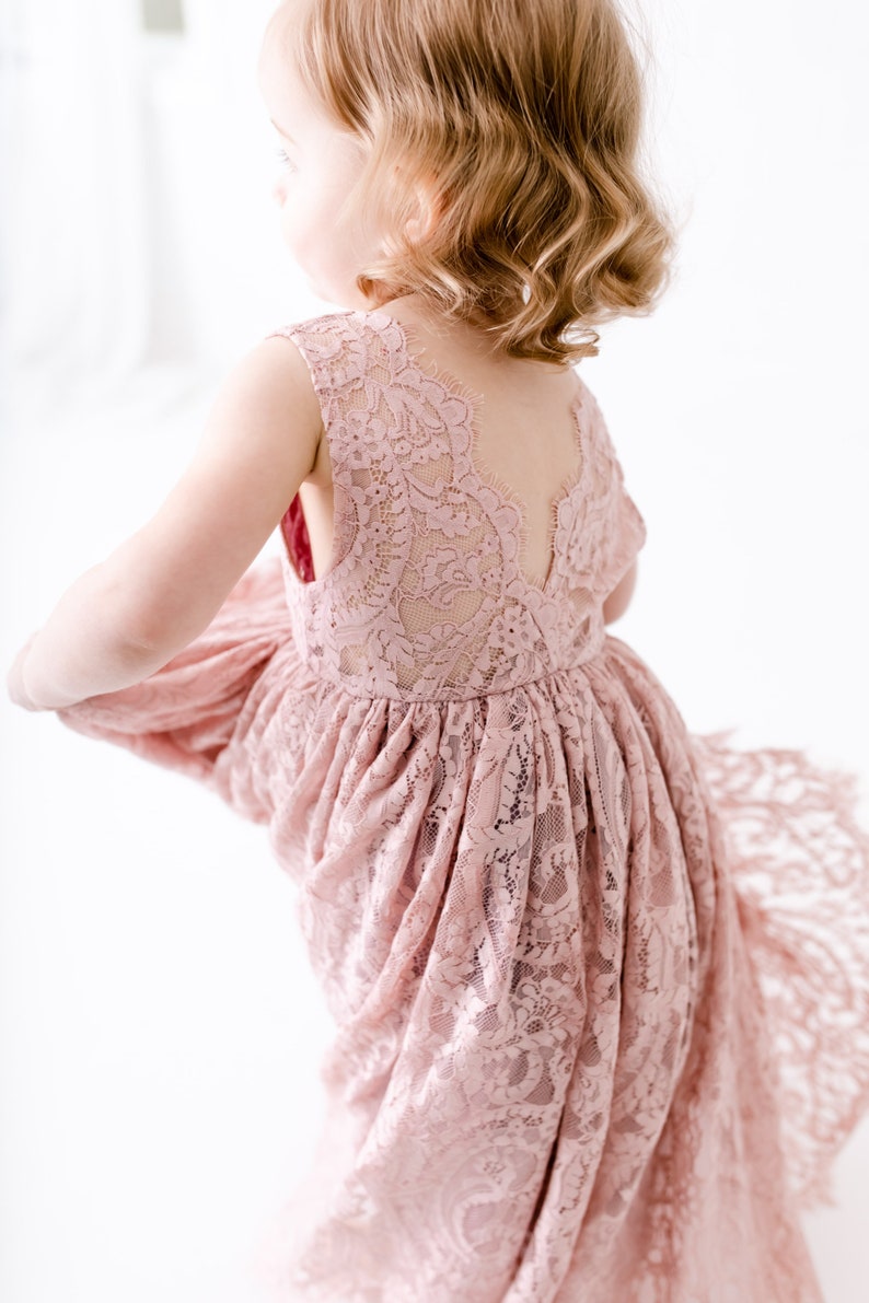 Pu&ograve; includere: Una bambina che indossa un abito di pizzo rosa polvere con schiena a V. L'abito ha una gonna fluida ed &egrave; perfetto per un'occasione speciale.