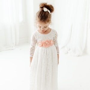 May include: A white lace dress with three-quarter length sleeves and a scoop neckline, featuring a waist sash adorned with three large, soft peach-colored fabric flowers. The dress has a full-length skirt with a scalloped hemline.