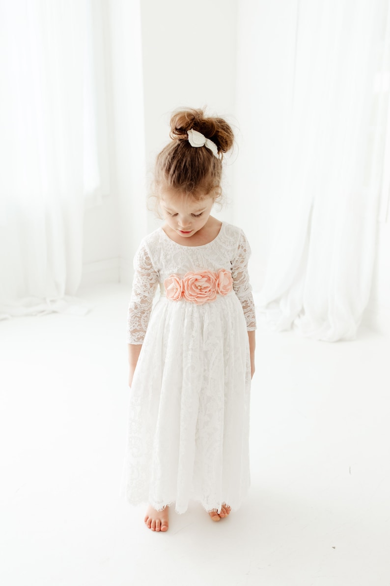 May include: A white lace dress with three-quarter length sleeves and a scoop neckline, featuring a waist sash adorned with three large, soft peach-colored fabric flowers. The dress has a full-length skirt with a scalloped hemline.