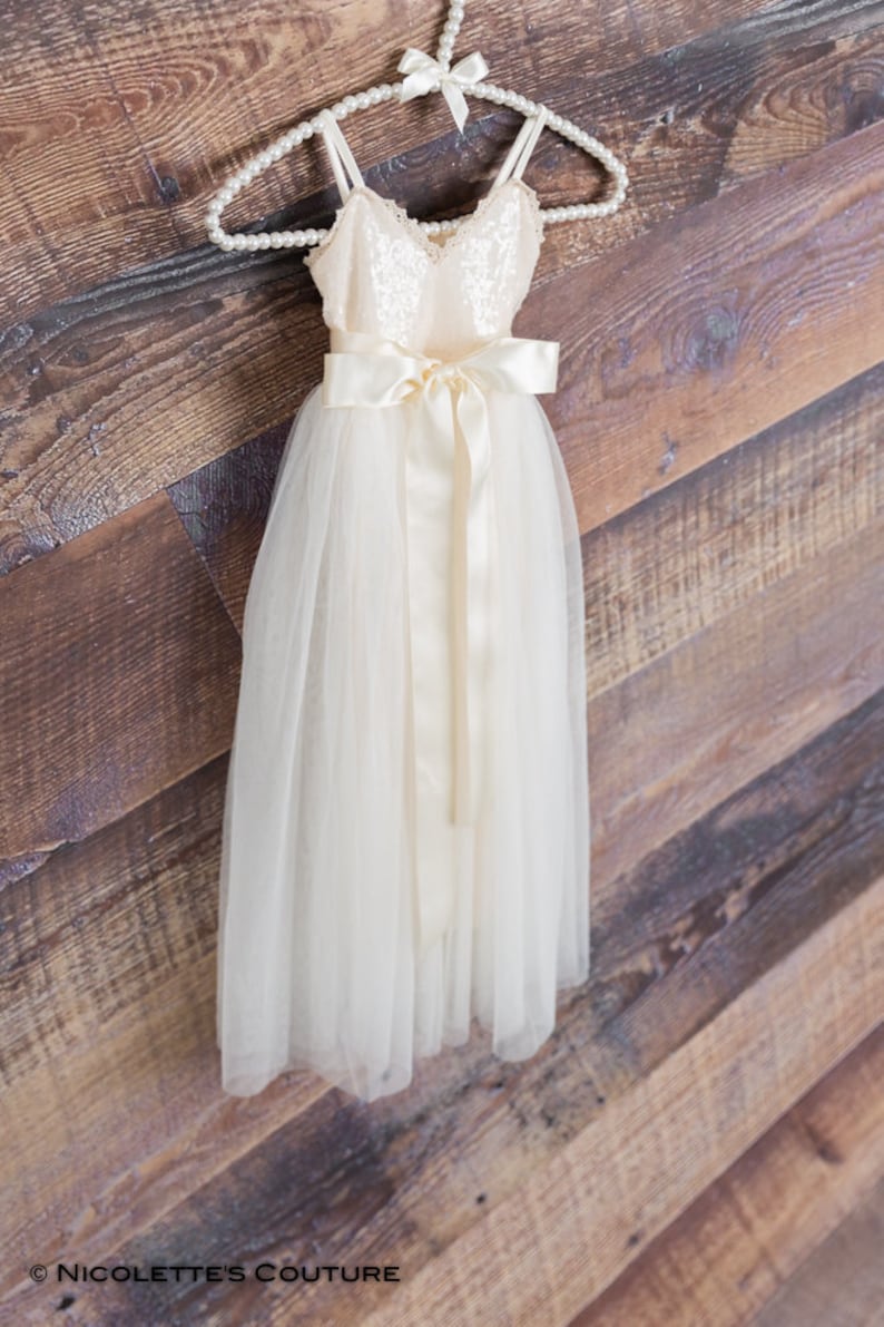 May include: A white tulle dress with a sequined bodice and a satin sash. The dress is hanging on a pearl-beaded hanger against a wooden background.