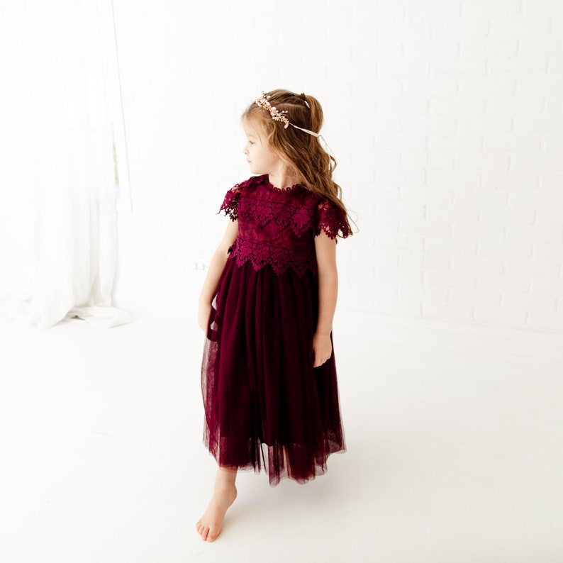 May include: A young girl wearing a burgundy tulle dress with a lace bodice and a gold headband. The dress has a scalloped hemline and the girl is standing on a white background.