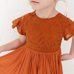 Op de afbeelding: Een jong meisje draagt een roestkleurige jurk met een kantboord en een gerimpelde zoom. De jurk heeft korte mouwen en een ronde hals. Het meisje draagt een bloemenkrone en heeft een serieuze uitdrukking op haar gezicht.