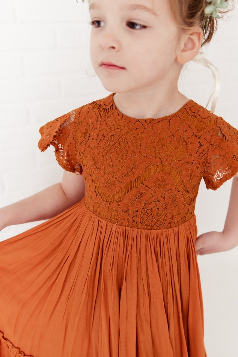 Op de afbeelding: Een jong meisje draagt een roestkleurige jurk met een kantboord en een gerimpelde zoom. De jurk heeft korte mouwen en een ronde hals. Het meisje draagt een bloemenkrone en heeft een serieuze uitdrukking op haar gezicht.