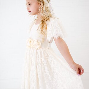 May include: An ivory lace dress featuring short flutter sleeves, a round neckline, and a satin waistband adorned with three fabric flowers. The dress has a scalloped hemline and is paired with a gold-toned floral headband with small white accents.