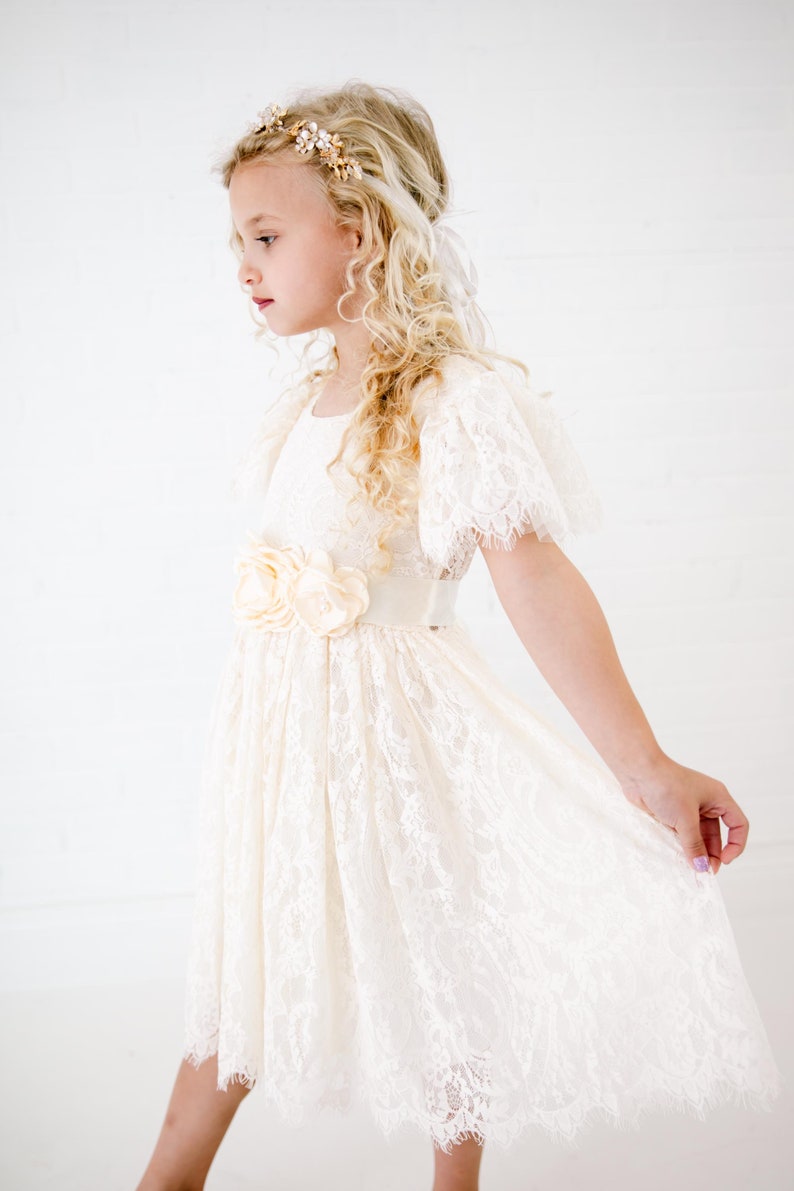 May include: An ivory lace dress featuring short flutter sleeves, a round neckline, and a satin waistband adorned with three fabric flowers. The dress has a scalloped hemline and is paired with a gold-toned floral headband with small white accents.