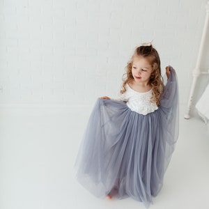 May include: A young girl wearing a white lace top with a grey tulle skirt. The skirt is flowing and the girl is holding it up with one hand.