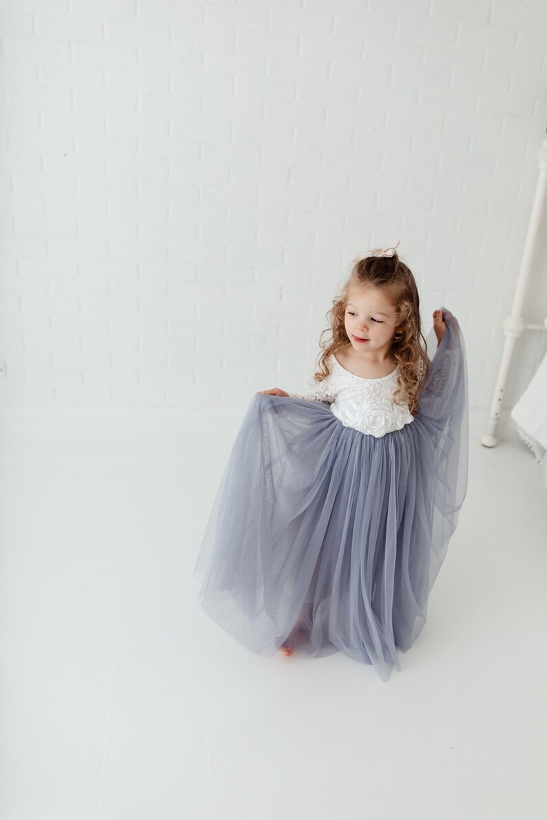 May include: A young girl wearing a white lace top with a grey tulle skirt. The skirt is flowing and the girl is holding it up with one hand.