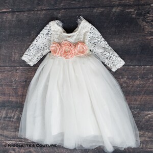 May include: A white tulle dress with long lace sleeves and a peach flower sash. The dress has a round neckline and a full skirt.