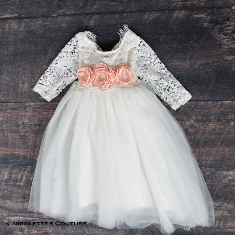 May include: A white tulle dress with long lace sleeves and a peach flower sash. The dress has a round neckline and a full skirt.