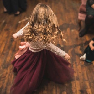 May include: A young girl in a white lace top and a burgundy tulle skirt dances on a wooden floor. Her hair is long and curly, and she is wearing a pink ribbon in her hair.
