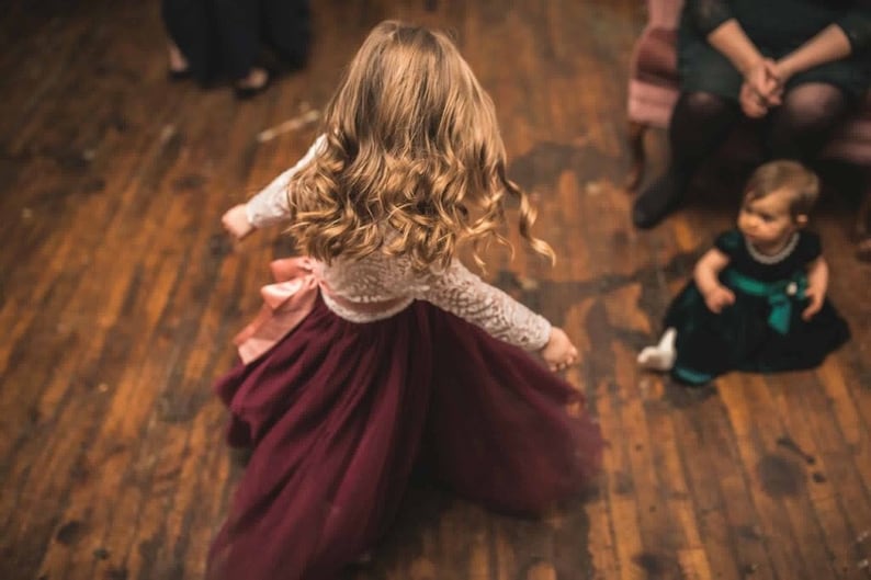 May include: A young girl in a white lace top and a burgundy tulle skirt dances on a wooden floor. Her hair is long and curly, and she is wearing a pink ribbon in her hair.