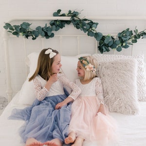 May include: Two young girls wearing white lace dresses with tulle skirts, one in blue and one in pink, are sitting on a white bed and smiling at each other. The bed has a white headboard with a garland of eucalyptus leaves.