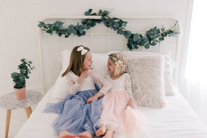 May include: Two young girls wearing white lace dresses with tulle skirts, one in blue and one in pink, are sitting on a white bed and smiling at each other. The bed has a white headboard with a garland of eucalyptus leaves.