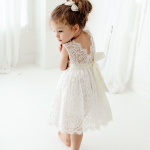 Pode incluir: Uma menina pequena vestindo um vestido branco de renda com uma faixa de cetim e um la&ccedil;o branco no cabelo. Ela est&aacute; em p&eacute; em um fundo branco.